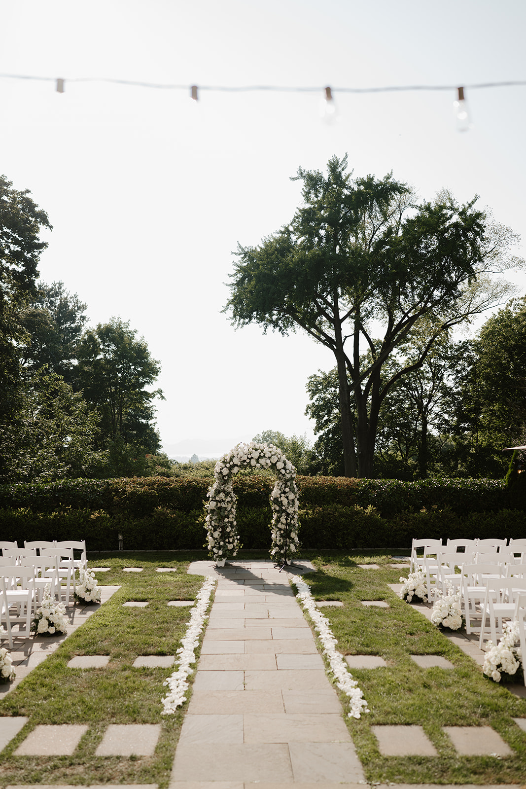 Outdoor wedding ceremony setup, featuring white chairs and a lush floral arch against green trees.